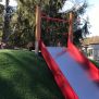 Hill Slide Wide SLW210, yapay çim kaplı tepe üzerine monte edilmiş kırmızı kenarlı metal kaydırak.